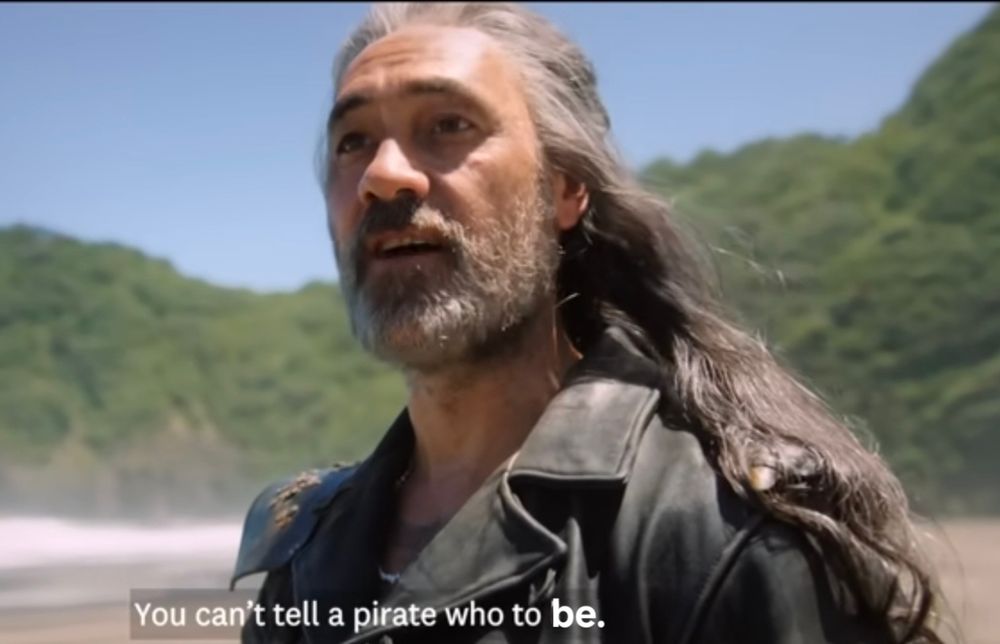 Taika Waitit dressed as Edward Teach from Our Flag Means Death. This screenshot is from a promotional video for visiting New Zealand. He's standing on a beautiful sunny beach, shown from the shoulders up in 3 quarter profile. The rolling green mountains and blue sky behind him are lovely. His beard is short, a season 2 look, and his long wavy hair flows behind his shoulders. He is saying, "You can't tell a pirate who to be." The original line was "love" instead of "be", but i think the sentiment is the same and we need to hear this right now.🏳️‍⚧️