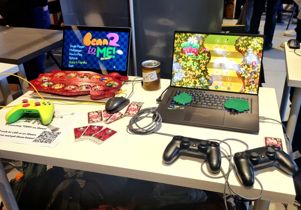 An event stand set up with two laptops and an assortment of game controllers. One screen displays the main menu of Bean To Me, and the other shows Springpongs menu.