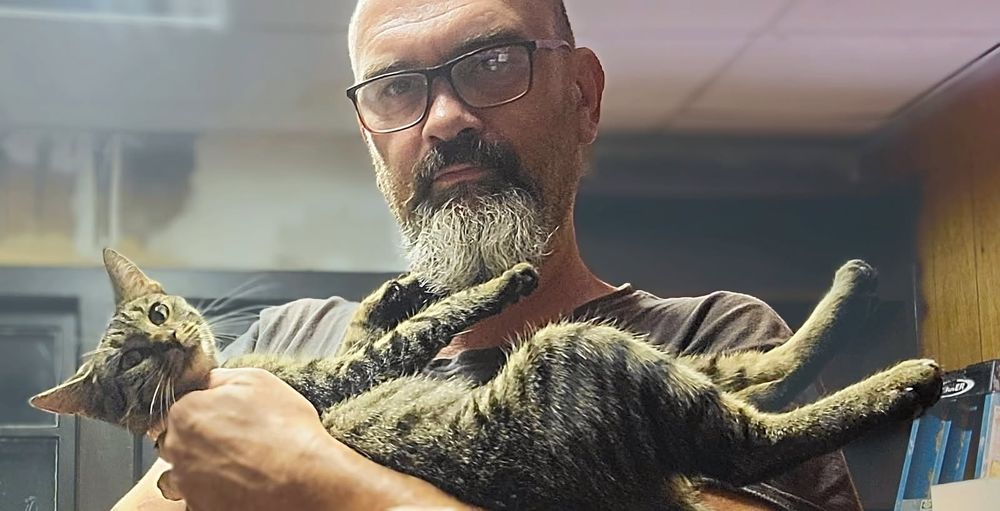 A scraggly bearded bald dude holding a gray tabby cat like a baby. Her legs are stretched out and both dude and cat are looking into the camera