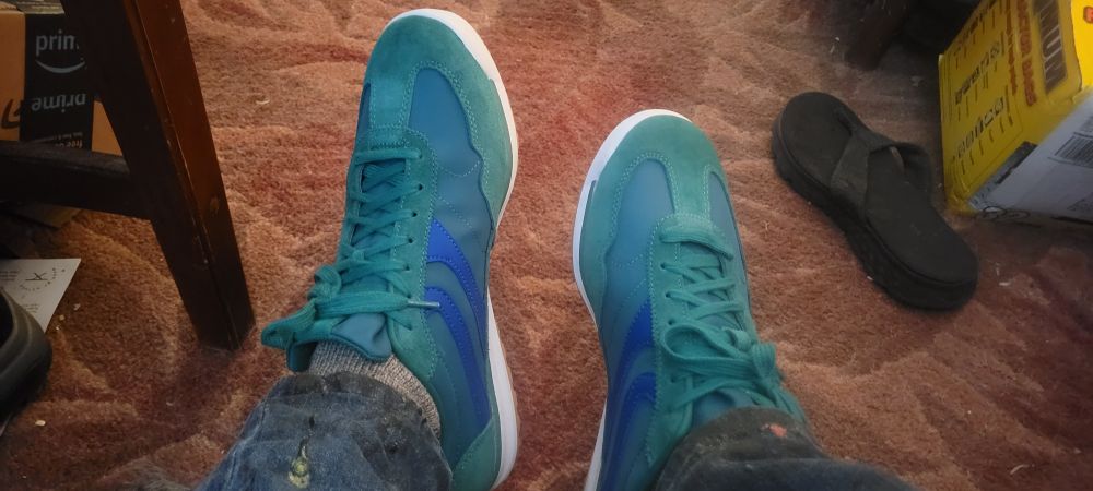 Green sneakers with white & gum colored soles. There's a table leg on the left, a single black flip-flop on the right, and it's all backed with a dirty rust-coloree carpet 