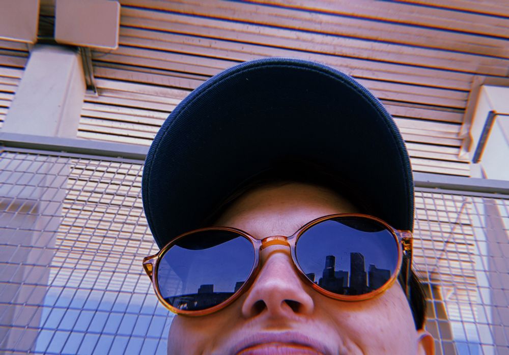 a selfie of just the top of my head from slightly below, im wearing a baseball cap and sunglasses and you can see the twin cities skyline reflected in the lens of one
