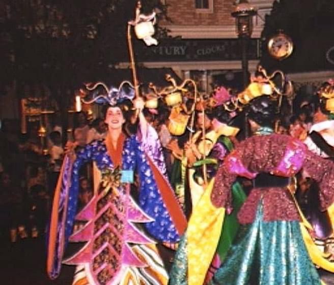 A group of Mulan Parade dancers on Disneyland Main Street at night. The costumes are multicolored silks, some with floor-length sleeves. The dancers have headpieces like black wigs with large flowers, and they are carrying lanterns on three-foot long poles. 