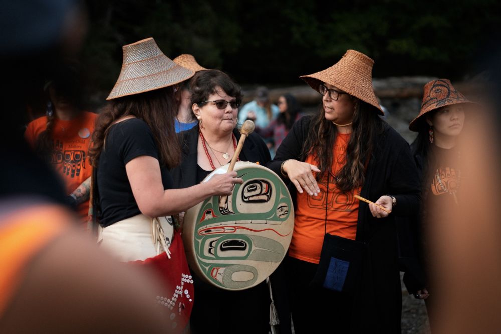 I am standing (left) with two of my clan sisters at our 2022 pole raising at Xaayna, Haida Gwaii. I am wearing a fancy woven cedar bark hat by Marlene Liddle, holding my gaawjaaw (drum) designed and painted by my brother Iljuuwaas Tyson Brown. The design on the drum is one of our crests, the maamats'iigaay (dragonfly).