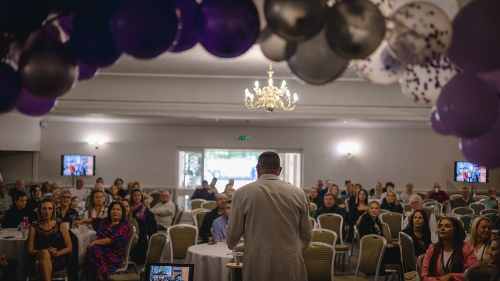 A man in a beige jacket addresses a large seated audience. The photo is taken from behind him, framed with purple balloons, showing the crowd listening attentively.