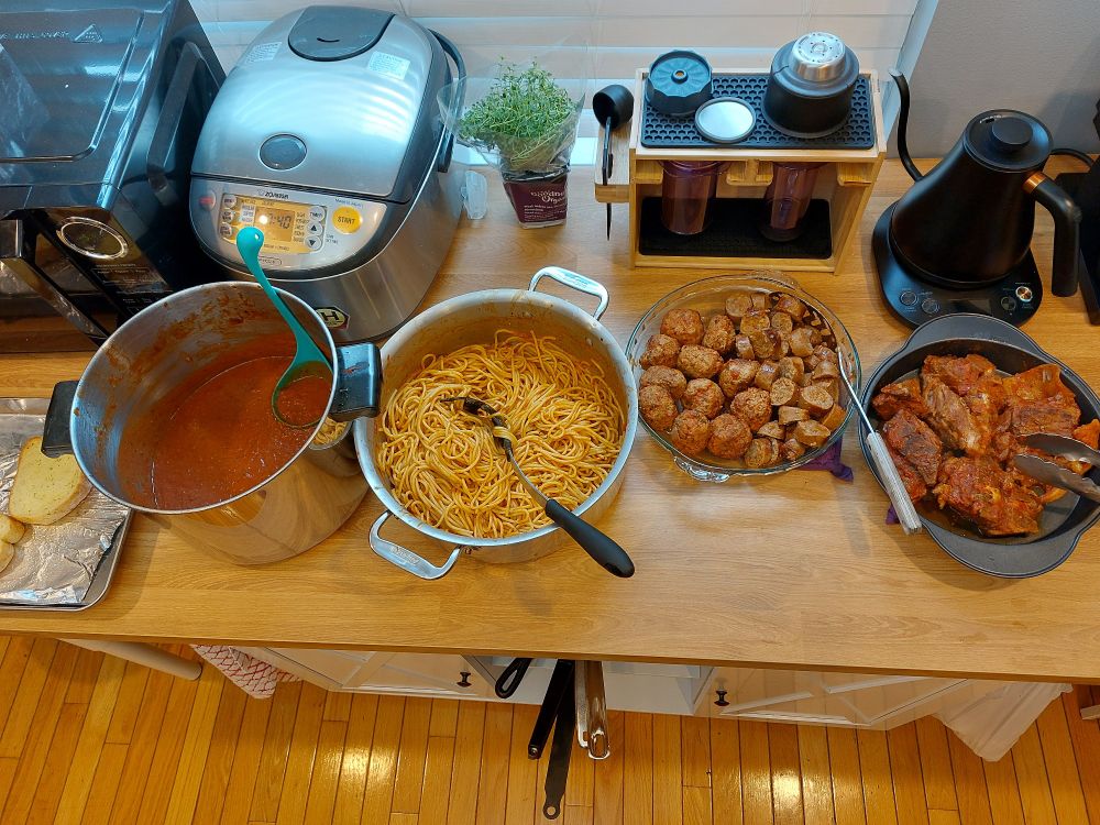 A wide shot of a counter, containing, left to right, garlic bread, a stock pot of Sunday sauce (Tomato), a pot of spaghetti, coated in sauce, a glass pie dish of meatballs and sausage (cooked in the sauce), and a cake tin of braised pork neck bones.

