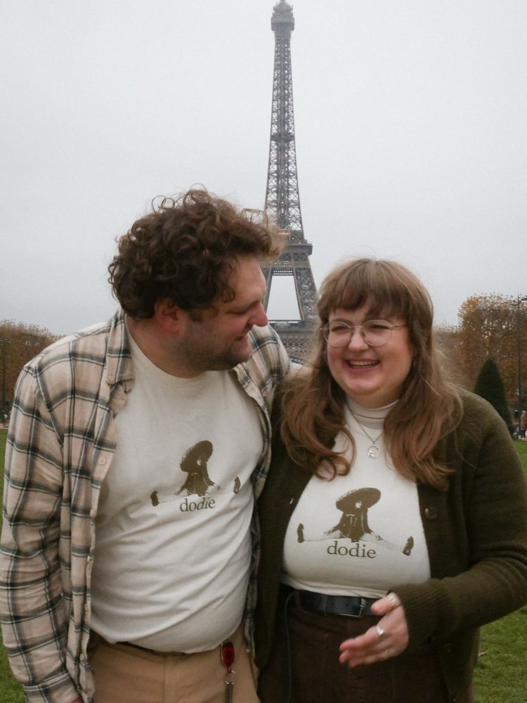 wouter looking at emily, both smiling, in front of the eiffel tower. they're both wearing a creme dodie tshirt with a picture of dodie on it. wouter is wearing a creme plaid flannel and light brown trousers. emily is wearing a green cardigan and brown skirt 