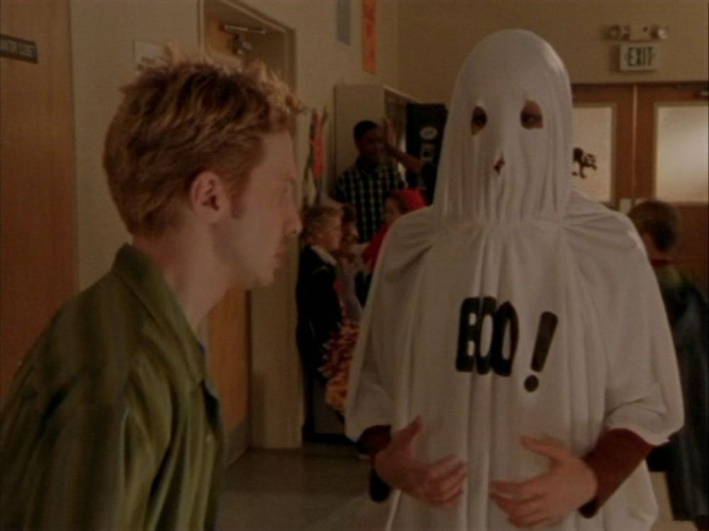 Willow trying to talk to Oz, from Buffy the Vampire Slayer. She is dressed in white sheet with “Boo!” written on it. He seems confused. 