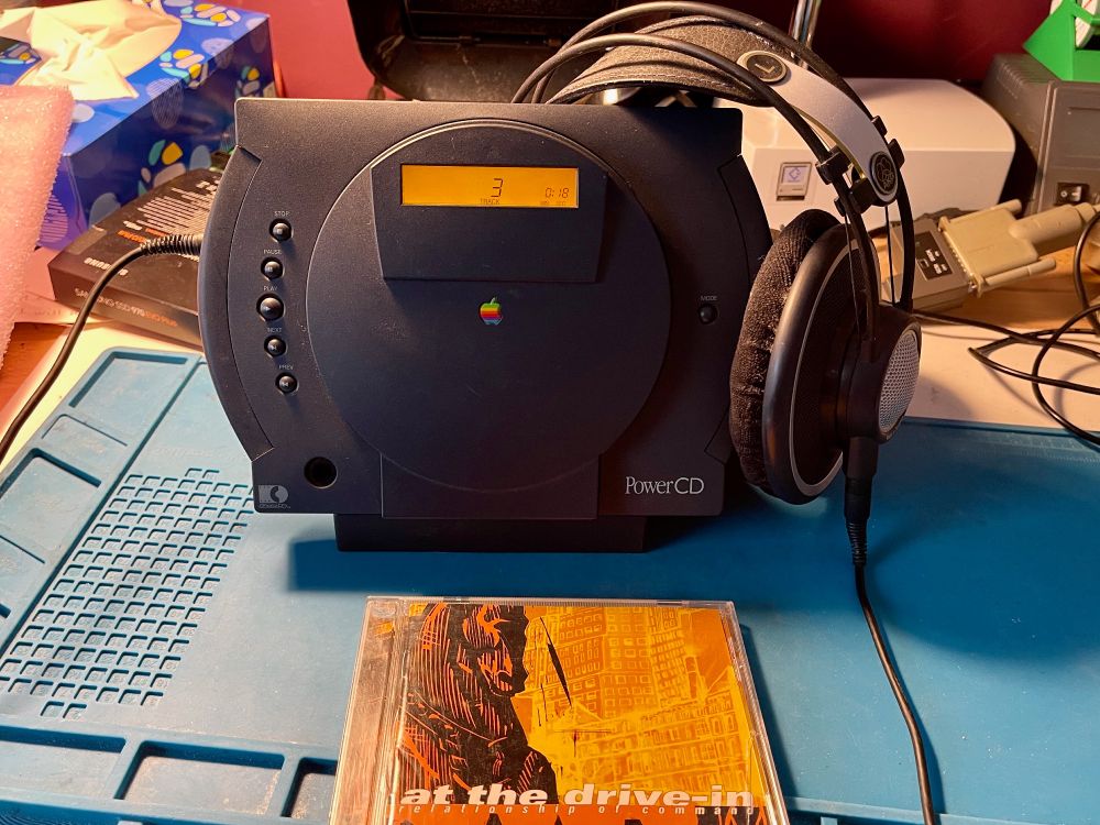 An Apple PowerCD CD player on a workbench with a pair of headphones and the box of the CD "Relationship of Command" by At the Drive-In.