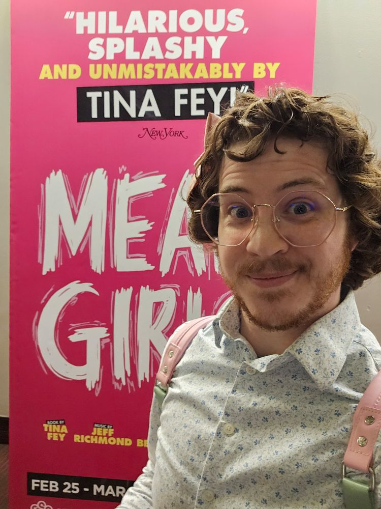 A young man with curly red brown hair and a white button down with a blue pattern smiles in front of a hot pink sign for the mean girls musical 