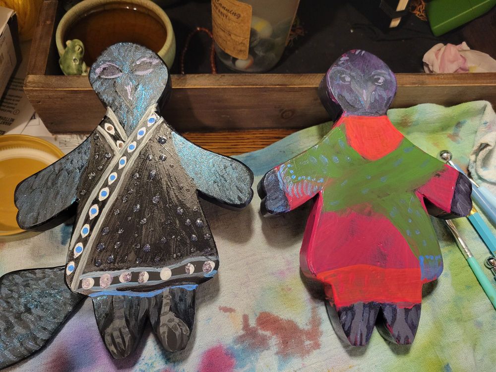 A photo of two vaguely humanoid-in-a-dress shaped pieces of wood, painted to look like anthropomorphic crows in little outfits. One is wearing a black wrap smock with eye trim. The other wears a magenta and orange smock with a green scarf. Both have a layer of iridescent glitter over the feathered sections. 