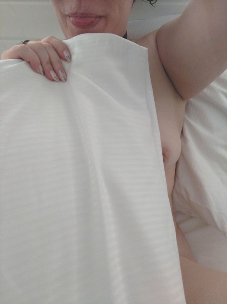 Frontalaufnahme von mir, ich sitze auf dem weiß bezogenen Bett, bin nackt, bedecke mich aber mit einem großen, weißen Kissen. Ich strecke die Zunge heraus, seitlich sieht man einen Teil meines Nippels neben dem Kissen hervor spitzeln. 