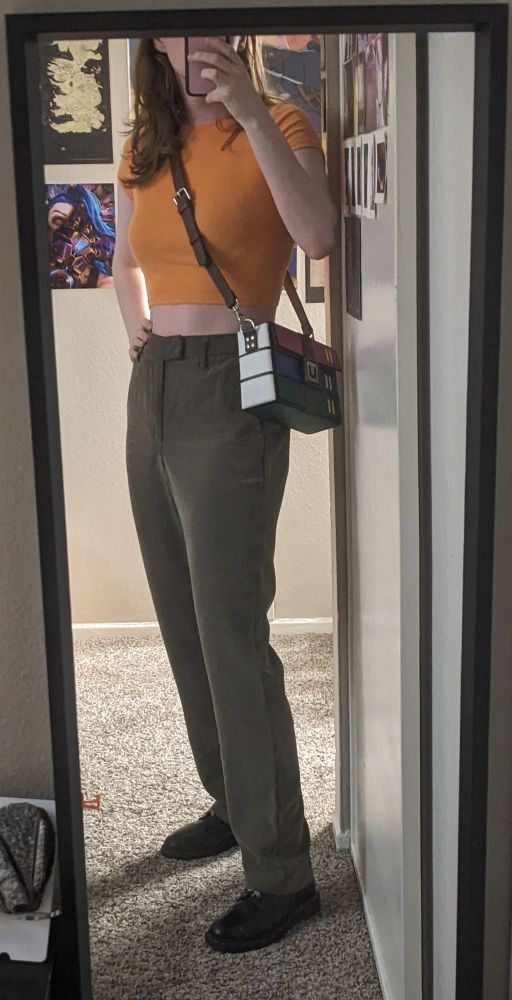 a stanced up transsexual wearing a tight fitting bright orange crop top, medium high waisted olive pants that are ironed, and dirty black doc martens. also winrar crossbody bag