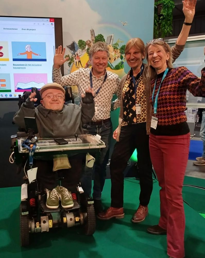 Een groep van vier mensen poseert samen op een groene vloer. Eén persoon zit in een elektrische rolstoel. De andere drie mensen staan naast de persoon in de rolstoel en hebben hun armen omhoog geheven. Op de achtergrond is een groot scherm te zien met kleurrijke illustratie. De sfeer lijkt vrolijk en ondersteunend, wat de inclusiviteit en samenwerking benadrukt.