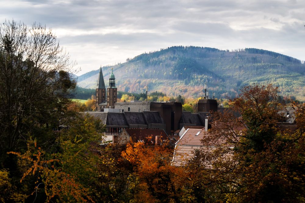 The view from the northern outskirts of the city of Goslar presents a picturesque scene with autumn-coloured trees in the foreground, alongside some rooftops of the town. The two towers of the Market Church stand out as a prominent focal point, with their copper roofs tinged with verdigris. In the background, one can observe the slope of the Steinberg, partially bathed in sunlight.