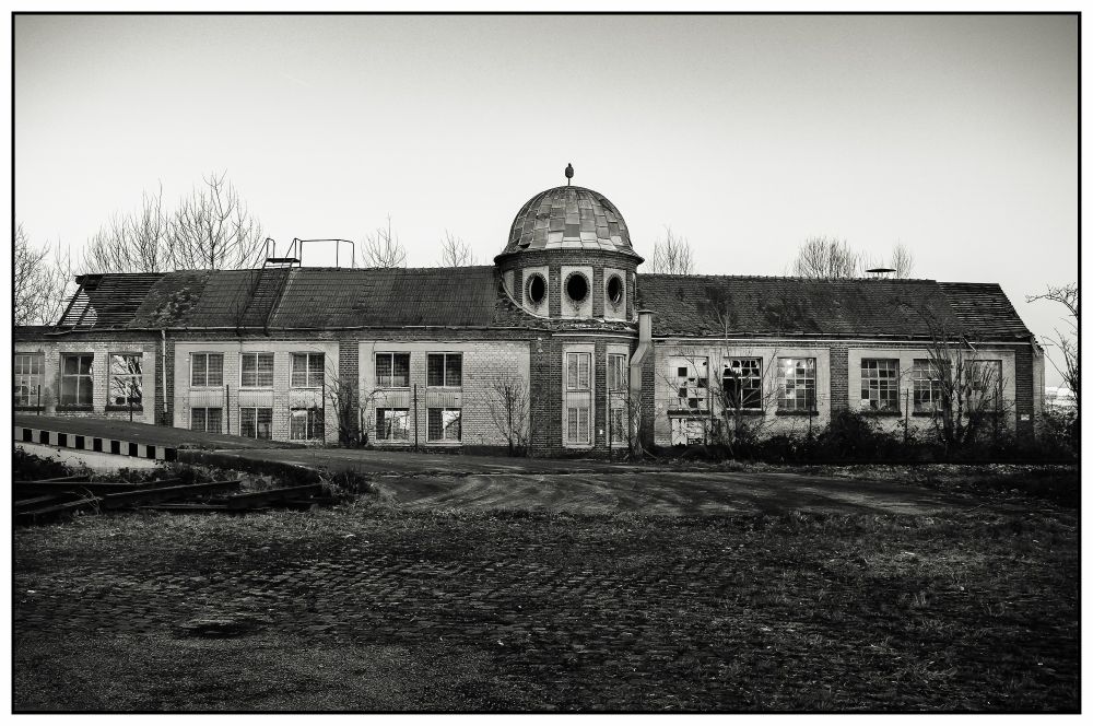 The photograph depicts the building of a former textile company. The elongated structure is captured in its entirety. A small turret in the centre visually divides the building into two halves. In reality, the turret is merely an architectural hint on the façade, a playful use of form. The photograph shows the state of the building long after the company ceased operations. The site has been cleared, and the actual production halls had already been demolished at the time the picture was taken. By January 2012, when the photo was taken, it was already decided that this building would also be demolished. It stands as a ruin: some window panes have been broken, and the roof shows signs of damage.