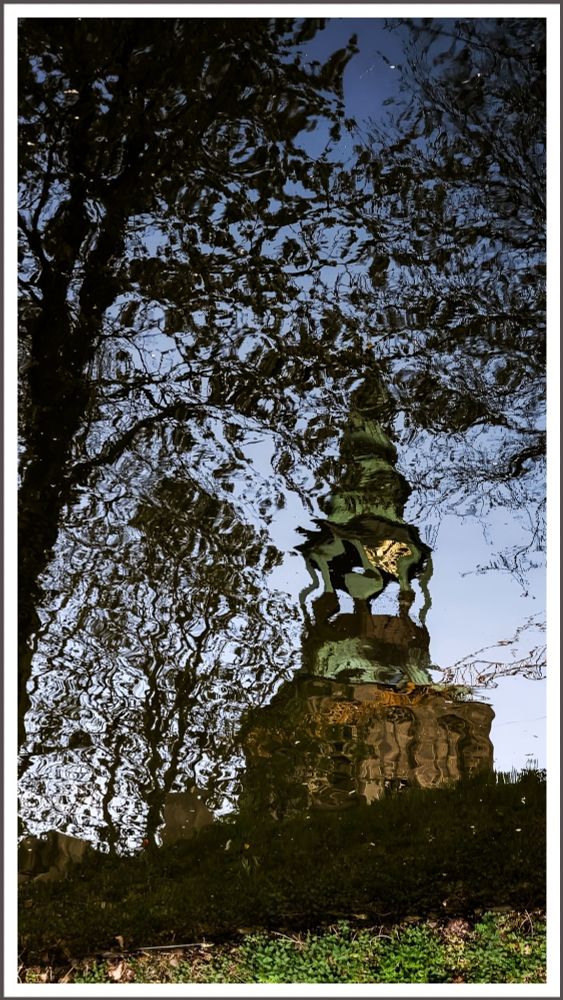 A church tower with a verdigris-covered spire and a tree are reflected in the water of a river. Their reflections are slightly distorted. At the bottom edge of the image, you can see the riverbank, which is covered with grass.