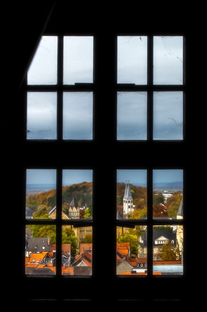 The camera gazes outward through the tower window of the Goslar Market Church. The window frame is entirely black. Through the panes, one can look down upon the northern part of the city, observing several red rooftops and a green hill in the background. The white twin tower of a church is visible, with the perspective creating the impression that the church has only a single tower. Above the scene, a rain-laden, dark blue sky looms.