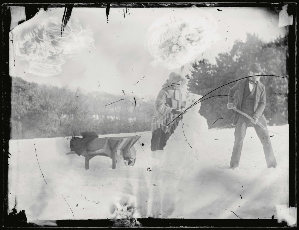 OLd black and white photo, two people making a snowman in mid-19th century clothes