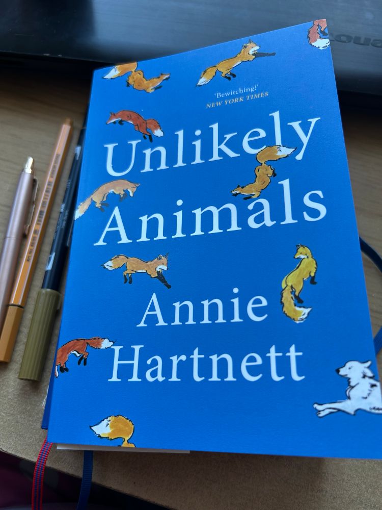 Book on top of a desk. Book cover is blue and dotted with small foxes. One small white fog in the bottom right hand corner. Book is called unlikely animals by Annie hartnett. Next to the book are three pens.
