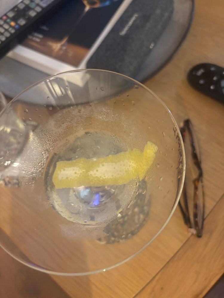 An overhead view of a martini glass a third full with lemon twist. General detritus on the table including reading glasses