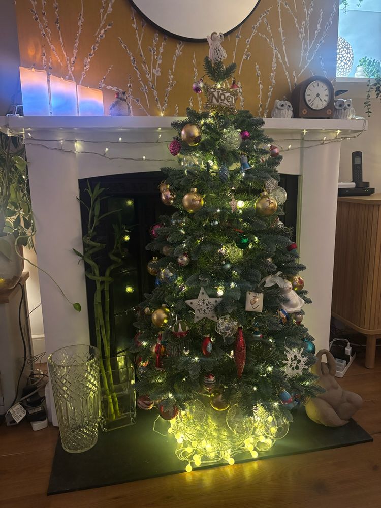 A Christmas tree with lights and baubles  placed in a fireplace hearth, with lights around the base. On the mantelpiece are 3 candles and a clock