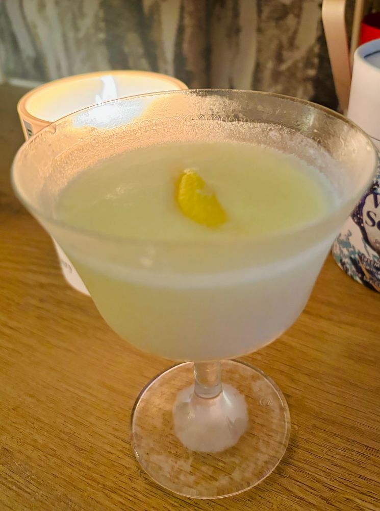 A glass on a wooden desk. The glass is filled with yellow liquid (gin, vermouth and lemon curd) and there’s a twist of lemon peel on top. Behind the glass is a lit candle 