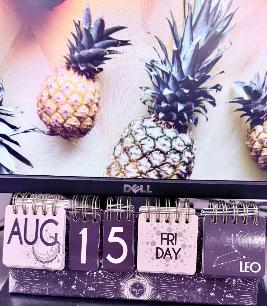 Desk calendar showing Friday, August 15th in Leo season, with a computer in the background showing a pineapple screen saver