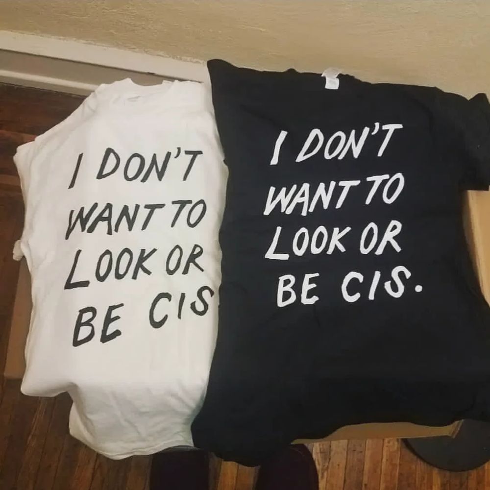 a white shirt w/ black text & black shirt w/ white text folded over a box, both reading in all-caps handwriting "I DON'T WANT TO LOOK OR BE CIS."