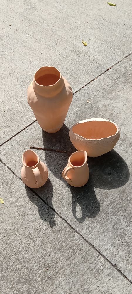 4 unglazed, bisque fired ceramic vessels with defects placed on the sidewalk (near a dumpster which is out of view). There are two small pitchers, a small football-shaped vase and a taller vase.