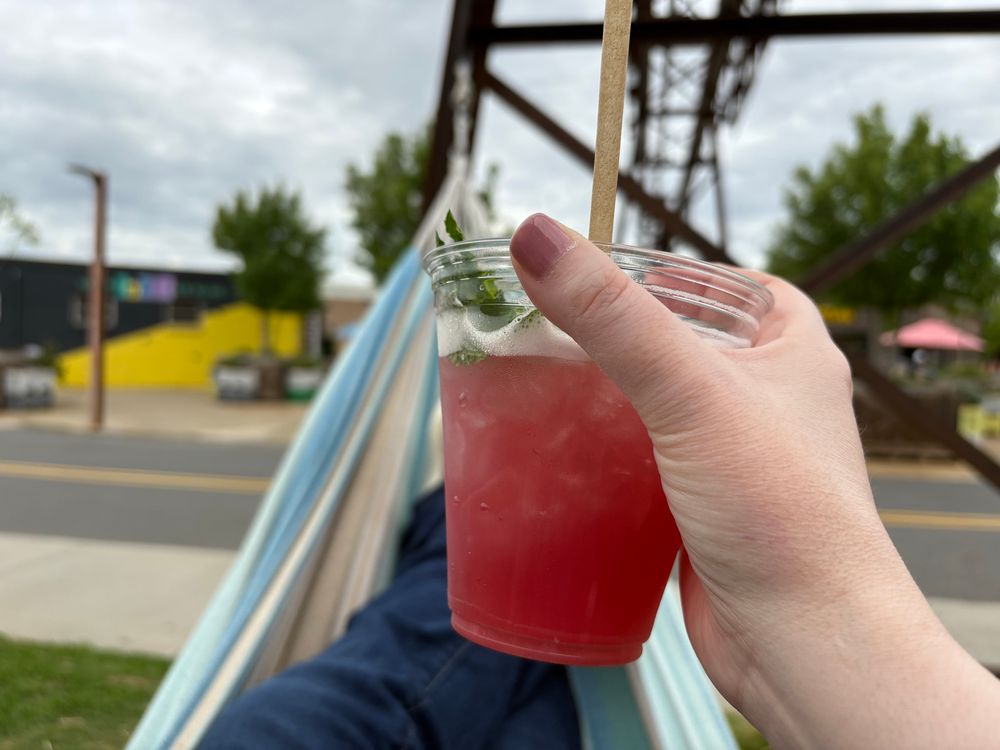 In the background there are several colorful buildings in Camp North End, with railroad infrastructure throughout. The forefront features a pink beverage and a very comfortable Mande in a hammock. 