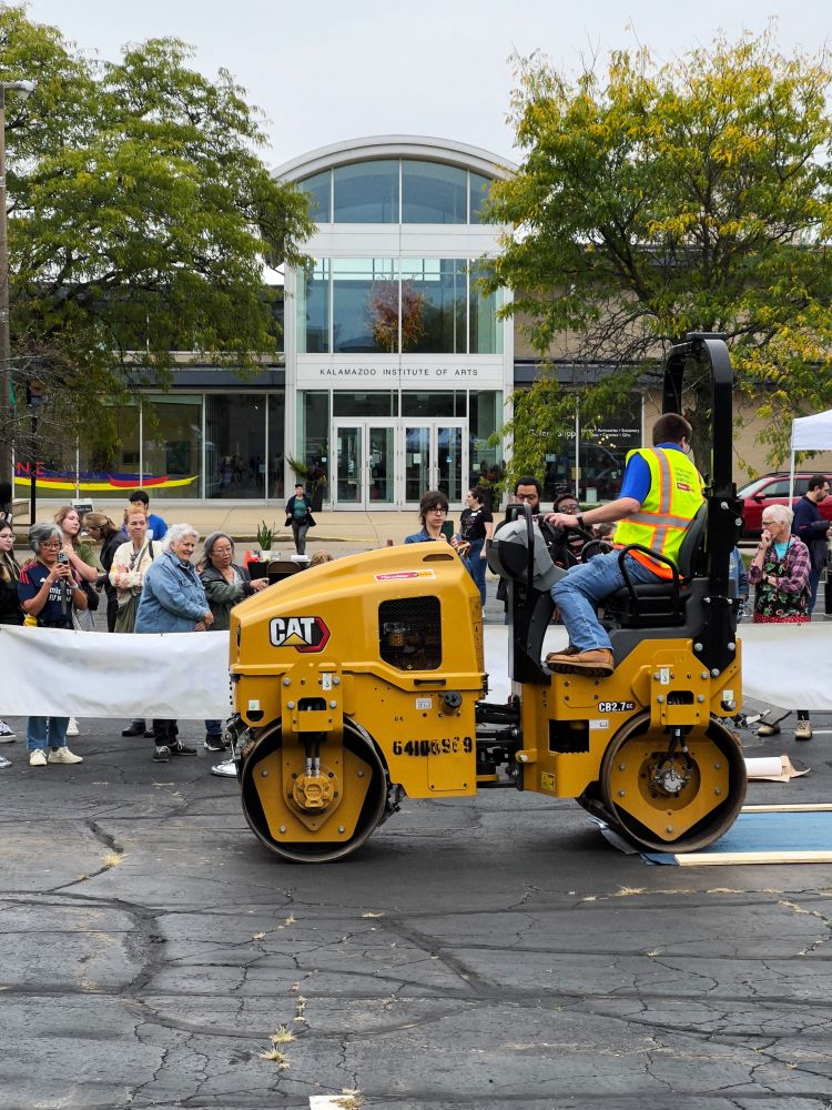 A steamroller drives over an inked woodblock in a parking lot in order to produce a canvas print as onlookers stand and watch from behind a barrier. The Kalamazoo Institute of Arts entrance is seen in the background.