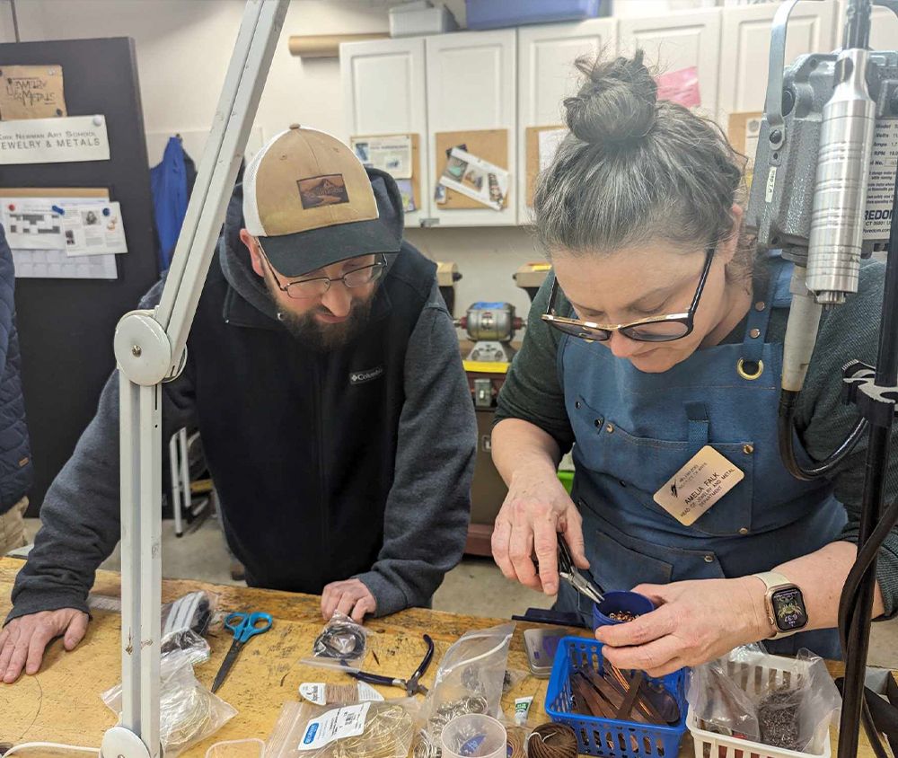 A person in a blue apron and KIA nametag uses needle-nose pliers to shape a piece of jewelry while a person in a gray sweatshirt and hat watches closely. They stand over a table with jewelry making supplies in bags and baskets.