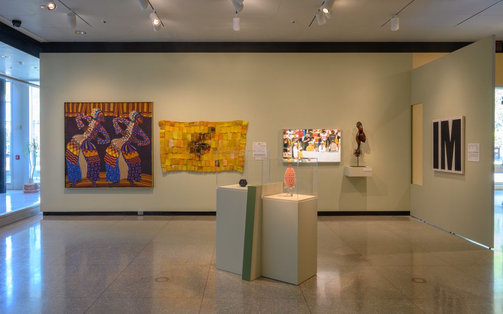 An art gallery with four artworks of different mediums hanging on a wall, and two others - including a black ash basket - on pedestals. Photo by Colleen Woolpert.