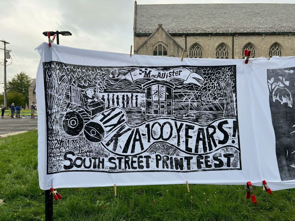 A black and white woodblock print celebrating the South Street Print Fest and the Kalamazoo Institute of Arts' 100th anniversary hangs with clothespins as it dries. The print itself depicts the event, showcasing a steamroller and the KIA building.