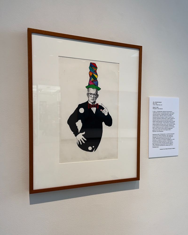 A photo of a framed lithograph of artist Kirk Newman. He wears a black suit and lifelike expression, humorously disrupted with a bright and multicolored geometric top hat and oversized striped bowtie. Circular cutouts in the suit and the exaggerated gesture of the subject's hand refer to the imagery seen in Newman’s own lithographs. 

Image credit: Jill Waskowsky, Kirk II, 1983, lithograph, hand-colored. Collection of the Kalamazoo Institute of Arts; Donation from Edward Sackley III, 2006.