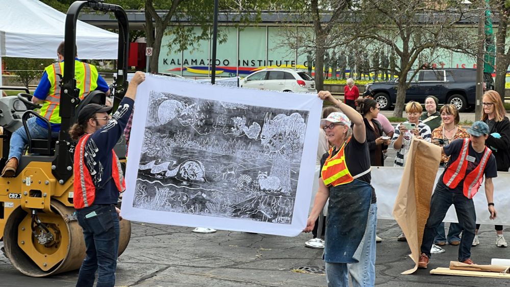 Two people wearing bright safety vests reveal a printed canvas produced by driving an asphalt roller over an inked woodblock. People watch and cheer for the reveal in the background from behind a barrier.