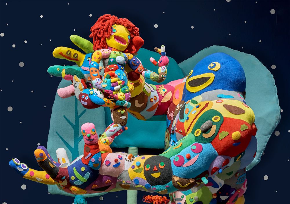 A photograph of a sculptural installation of comic figures, each containing dozens of panels of colorful fabric with shapes resembling faces. The larger figure is holding a smaller one aloft in one of its hands. They stand against a backdrop of trees and a starry night sky.