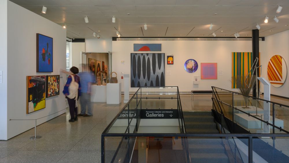 An art gallery with numerous abstract artworks on a wall in groupings and pairings, with a variety of bright geometric shapes and patterns. The gallery space surrounds a staircase to a lower gallery. Two visitors are shown in a motion blur. Photo by Colleen Woolpert.