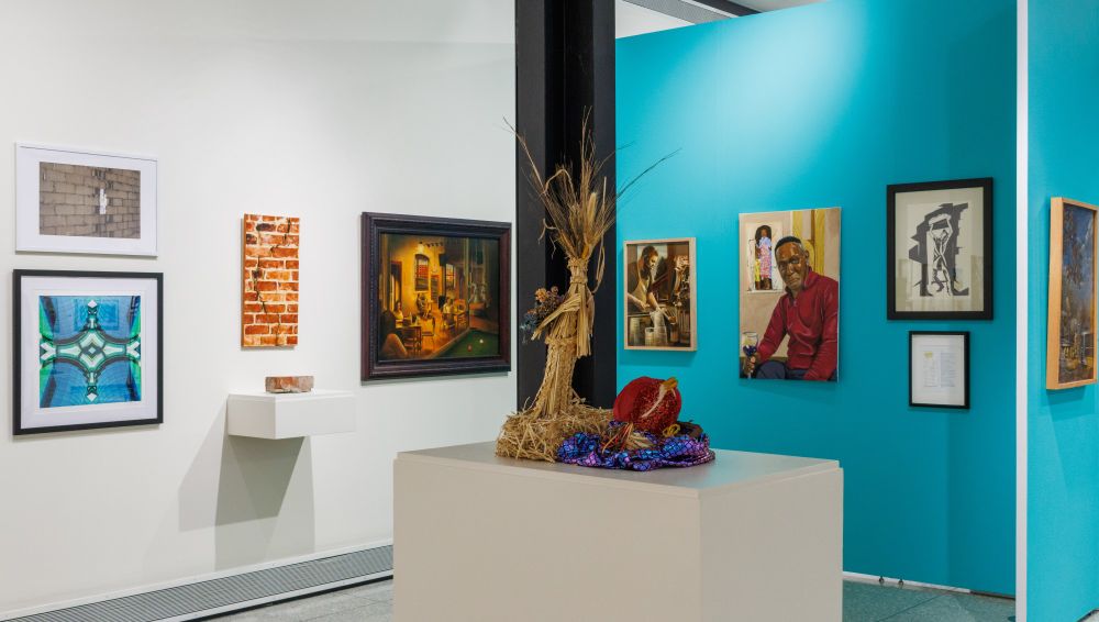 Art, including paintings and portraits, hangs on two gallery walls - one blue and one white - while a sculptural installation sits on a pedestal in the foreground.