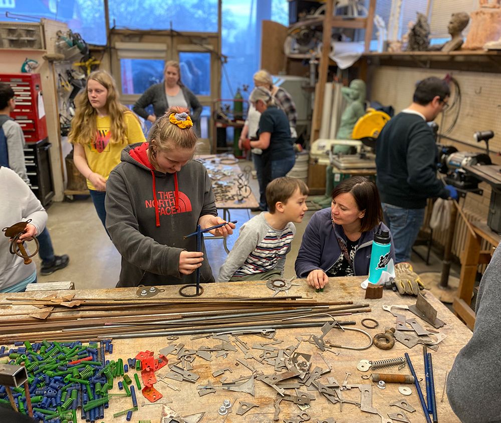 Children and adults work on assembling metal pieces into larger sculptures at different tables in a large metalworking studio.