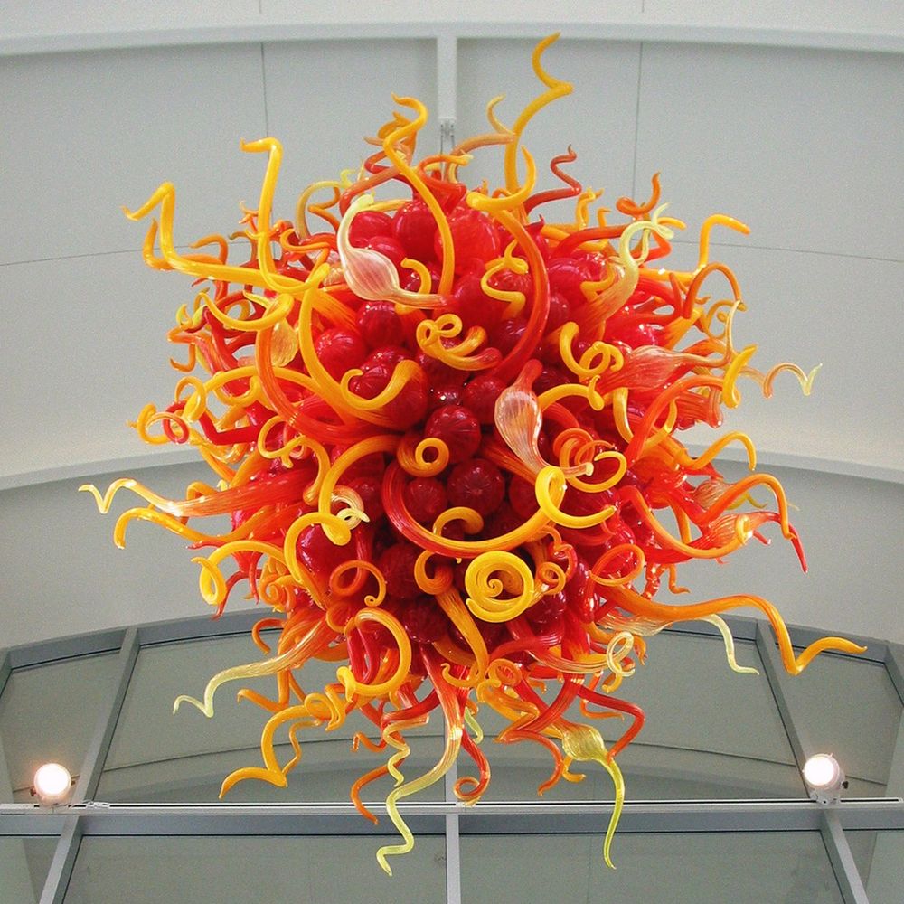 A hand blown glass sculpture hangs from a ceiling, featuring hundreds of red, orange, and yellow twisting and spiraling glass tubes and shapes radiating out from the center.