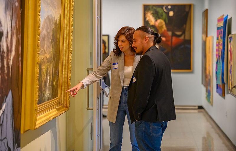 Two people, each wearing a blazer and name tag, stand in an art gallery as they point at and discuss a painting in a large and ornate gold colored frame. Photo by Colleen Woolpert.