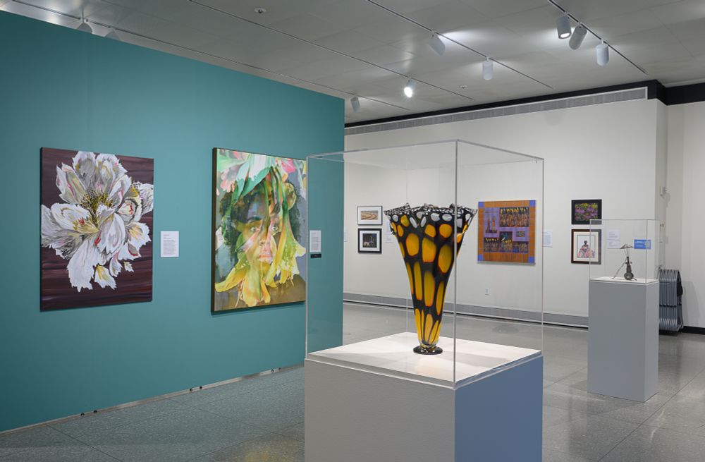 A glass sculpture with a color pattern resembling a monarch butterfly wing sits in a display case in front of a wall of several paintings at the Kalamazoo Institute of Arts
