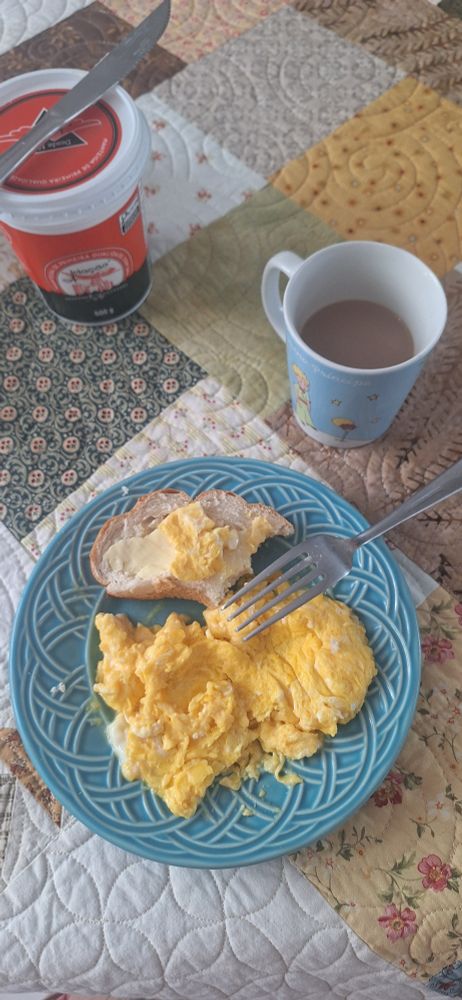 Pão de leite mordido num prato azul acompanhando ovo mexido e um café com leite.