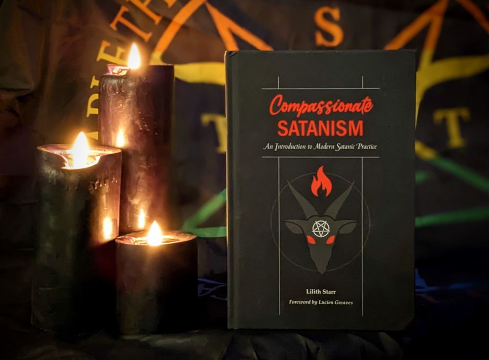 A copy of the book "Compassionate Satanism" by Lilith Starr standing in front of a Satanic Pride flag from the Satanic Temple.