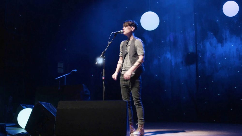 the poet Andrea Gibson on stage, standing in front of a microphone, performing. The backdrop looks like it has stars, and there are glowing orbs on the ground. Andrea wears a vest, and looks triumphant.