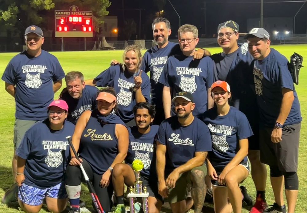 A softball team posing with a big trophy on a large field. They're smiling, and in the background is a scoreboard showing the visitors with 2 runs, and the home team with 12 runs.