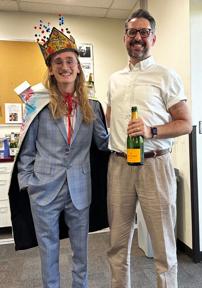 Two people standing next to each other. I'm on the right, wearing a white shirt and holding a bottle of celebratory champagne. Jake, who was just awarded his PhD, is on the left in a blue suit and his cape and crown. The crown is painted gold, and has little puff balls above representing galaxies in the early Universe. 