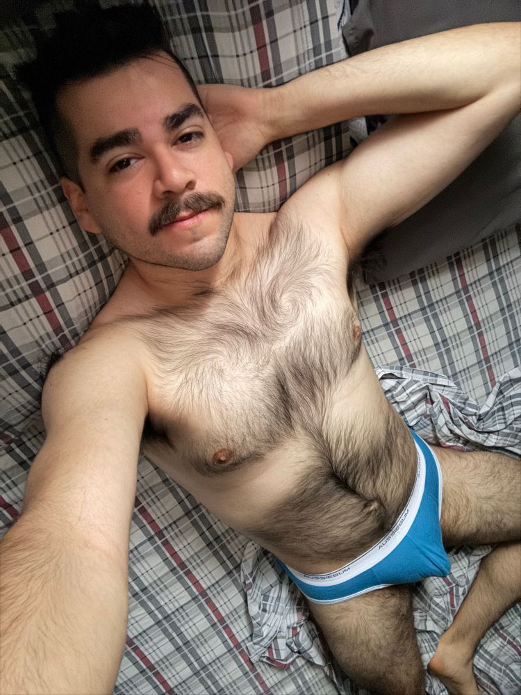 Selfie of man in bed wearing blue briefs, has one hand behind his head