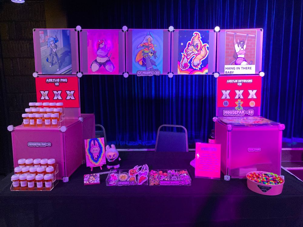 A dealer’s den table set up. Prints are on top, on the table itself is stickers and button badges 